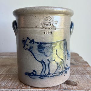 May include: A cylindrical stoneware crock with a light tan glaze and two side handles. The front features a blue hand-painted cow illustration and an impressed stamp reading HANDMADE ROWE POTTERY WORKS CAMBRIDGE WI 1990.