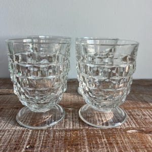 May include: Four clear, vintage-style drinking glasses. Each glass features a textured, geometric pattern around the body and sits on a small, round base. The glasses are made of clear glass and are displayed on a wooden surface.