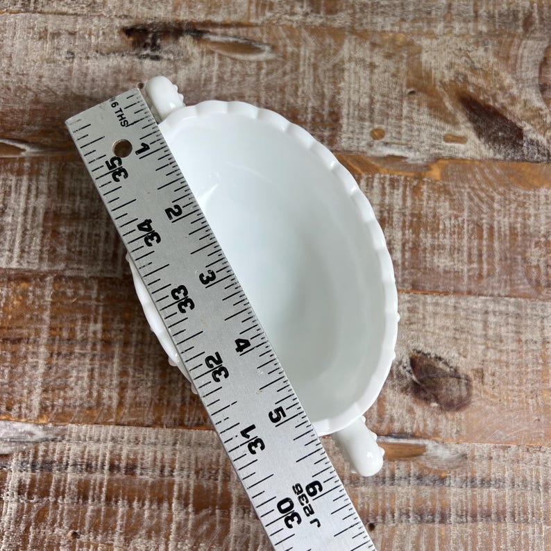 May include: A white milk glass bowl with a scalloped rim and two small handles, positioned beneath a metal ruler that shows the bowl measures approximately 5.5 inches in length from handle to handle and 3.5 inches in width at the center.