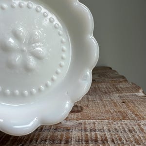 May include: A white milk glass plate featuring a scalloped edge and a central embossed floral design surrounded by a raised beaded circle. The plate has a smooth, glossy finish and a circular shape with a distinct decorative rim pattern.