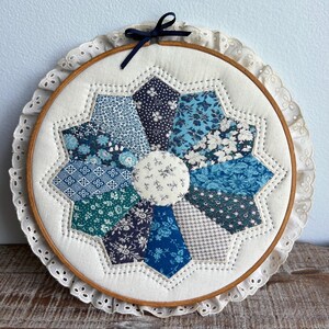 May include: A circular wooden embroidery hoop containing a fabric quilt block with a central white floral circle surrounded by ten radiating petals in various shades of blue and white patterns, finished with a white eyelet lace trim and a small black bow.