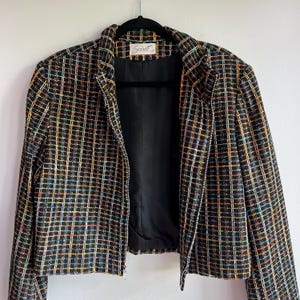 Op de afbeelding: Een zwart tweed jasje met een veelkleurig ruitpatroon van oranje, blauwe en gele draden. Het jasje heeft een zilverkleurige ritssluiting over de gehele lengte, een hoge kraag en een zwarte voering. Het label in de nek vermeldt de naam Soirée.