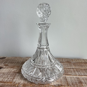 May include: A clear crystal decanter with a diamond pattern and a round stopper. The decanter has a wide base that tapers to a narrow neck and a decorative stopper. The glass is cut with a geometric design.
