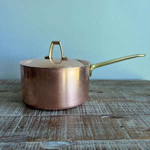 May include: A polished copper saucepan with a matching lid and a long, slender brass handle. The pot features a cylindrical body with a smooth, metallic finish and a triangular brass knob centered on the lid for easy lifting and handling.