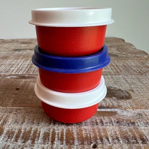May include: A stack of three small, round, red plastic containers with textured sides. The top and bottom containers feature white lids, while the middle container has a blue lid. Each container has a wide, flat rim extending from the top edge.