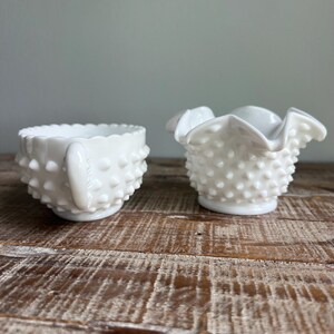 May include: A set of two white milk glass pieces featuring a hobnail texture. One piece is a small creamer with a handle and a scalloped rim, while the other is a small bowl with a ruffled, wavy edge. Both items have a glossy, opaque finish.