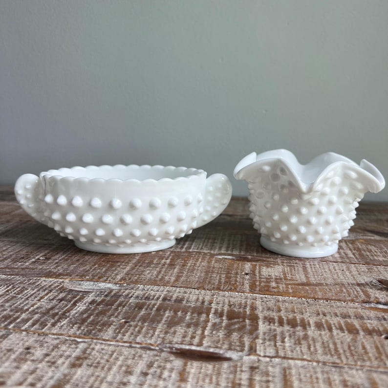 May include: A white milk glass bowl with two handles and a hobnail texture alongside a matching white milk glass creamer pitcher featuring a ruffled rim and a consistent hobnail pattern across the exterior surface of both glass pieces.
