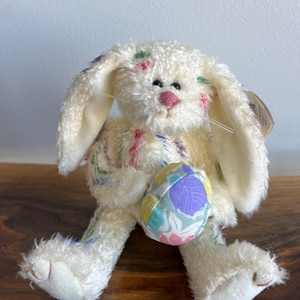 May include: A cream-coloured plush bunny with long ears and colourful embroidered accents. The bunny is holding a patterned Easter egg. The bunny has a pink nose and is sitting on a wooden surface.