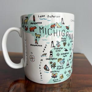May include: White ceramic mug featuring a map of Michigan with illustrations of landmarks and animals. The mug includes the text "Michigan" and various city names. The handle is white and the mug sits on a wooden surface.