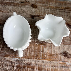 May include: Two white milk glass vessels, one oval-shaped with a scalloped rim and a small handle, and one star-shaped bowl with ruffled edges. Both pieces feature a smooth, glossy finish and are presented from a top-down perspective.