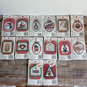 May include: A collection of fourteen individual Christmas ornament cross-stitch kits by The Needlecraft Shop. Each kit is packaged in a clear plastic sleeve with a decorative card backing featuring a unique holiday design, including Santa, animals, and festive text.