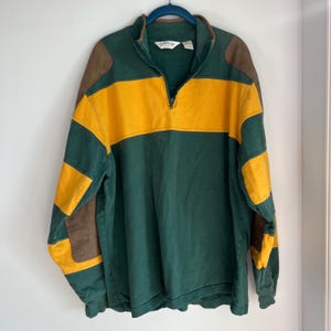 Peut inclure: Un pull à manches longues avec fermeture éclair au quart, présentant un design à blocs de couleurs vert foncé, jaune moutarde et marron. Le vêtement comprend des empiècements marron aux épaules, un col montant et une étiquette Orvis au col.