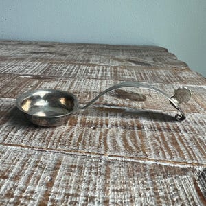 May include: A silver-toned metal ladle with a shallow, circular bowl and a long, curved handle. The handle features a decorative, scalloped-edge finial at the end, which curls slightly upward, providing a distinct profile for this serving utensil.