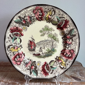 May include: A round ceramic plate featuring a central illustration of a harbor with a red-sailed ship and small boats, surrounded by a wide border of colorful pink, red, and yellow flowers with green leaves on a cream-colored background.