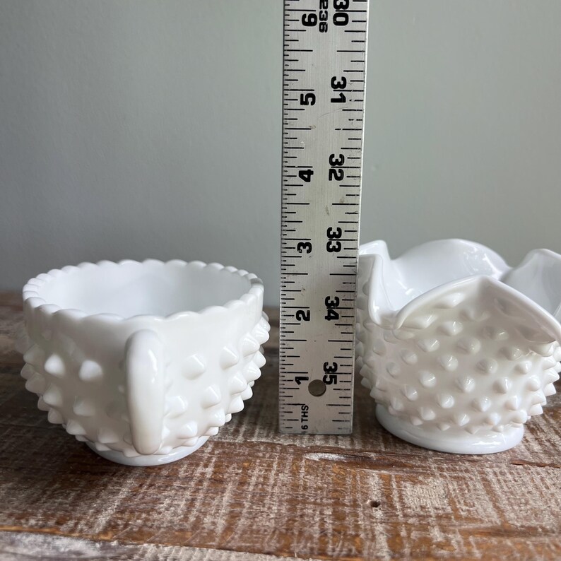 May include: Two white milk glass bowls with a hobnail texture. The left bowl features a handle and a scalloped rim, while the right bowl has a ruffled edge. A vertical ruler is placed between them, showing a height of approximately 2.5 inches.