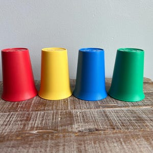 Könnte beinhalten: Vier bunte Plastikbecher in Rot, Gelb, Blau und Grün. Die Becher sind in einer Reihe auf einer Holzoberfläche angeordnet. Jeder Becher hat einen leicht ausgestellten Boden und eine flache Oberseite.