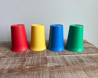 May include: Four colorful plastic cups in red, yellow, blue, and green. The cups are arranged in a row on a wooden surface. Each cup has a slightly flared base and a flat top.