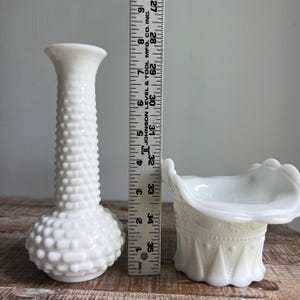 May include: A white milk glass bud vase with a hobnail texture and a white milk glass basket with a ruffled rim and embossed geometric patterns, positioned next to a ruler showing a height of approximately 9.5 inches for the taller vase.