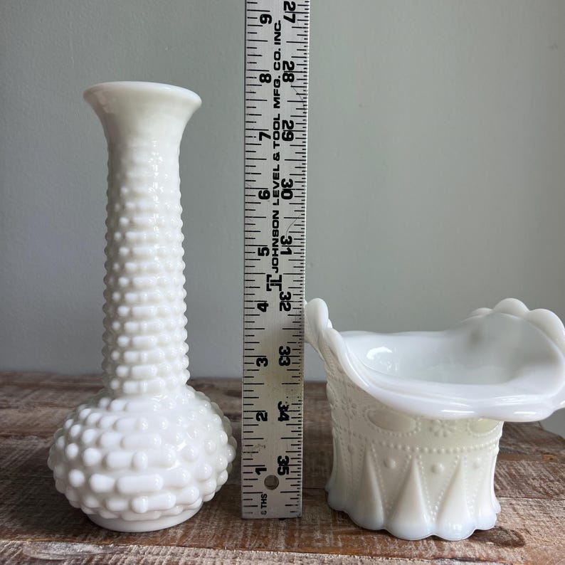 May include: A white milk glass bud vase with a hobnail texture and a white milk glass basket with a ruffled rim and embossed geometric patterns, positioned next to a ruler showing a height of approximately 9.5 inches for the taller vase.