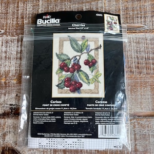 May include: A Bucilla counted cross stitch kit titled Cherries in a clear plastic bag. The package features a printed image of red cherries on a branch with green leaves and a dragonfly, with a stitched size of 4.5 inches by 6.5 inches.