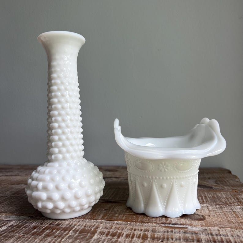 May include: A tall white milk glass vase with a hobnail texture and a shorter white milk glass bowl featuring a ruffled top edge and a decorative embossed pattern of dots and triangles around the exterior surface of the vessel.