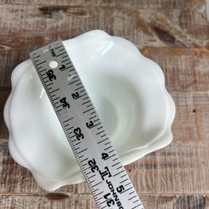 May include: A white milk glass bowl with a scalloped, petal-like rim. A metal ruler is placed across the bowl, showing a diameter of approximately 4.5 inches. The bowl has a smooth, glossy finish and a deep, rounded interior basin.