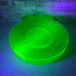 May include: A stack of bright green, circular glass plates. The plates are illuminated with a vibrant green light, creating a glowing effect. The plates are stacked neatly, showcasing their circular shape and smooth surface. The background features a textured brick wall.