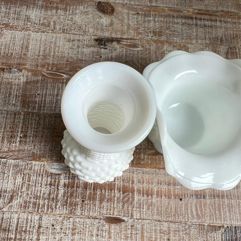 May include: Two pieces of white milk glass. One piece is a small vase with a hobnail texture and a flared rim. The second piece is a small pitcher or creamer with a smooth, ruffled, and scalloped edge opening. Both items are opaque white glass.