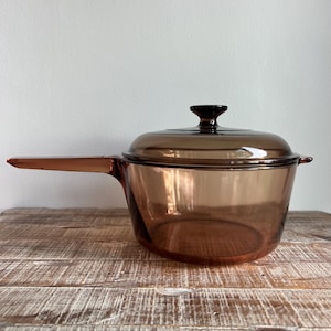 May include: A translucent amber-coloured glass saucepan with a matching lid. The cookware features a long, integrated glass handle on one side and a small tab handle on the opposite side, with a circular knob centred on the lid.