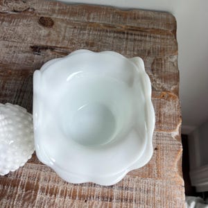 May include: A white milk glass bowl with a scalloped rim and a separate matching lid featuring a raised hobnail texture. The bowl has a smooth interior and a wide opening, while the lid is domed and covered in small, uniform rounded bumps.