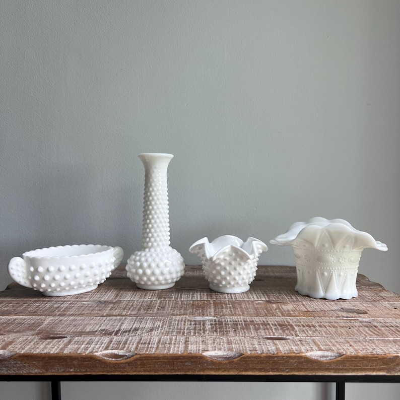 May include: A collection of four white milk glass vessels including an oval bowl with side handles, a tall bud vase with a hobnail texture, a small ruffled bowl with a hobnail pattern, and a wide vase featuring a scalloped rim and geometric details.