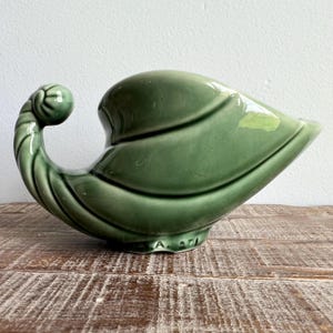 May include: A green ceramic leaf-shaped decorative vase. The vase has a curled handle and a wide, open mouth. The surface is glossy and smooth, with visible leaf vein details. The base has the markings "A. 271".