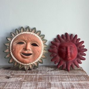 Könnte beinhalten: Zwei sonnenförmige Wandplatten. Die größere Platte ist terrakottafarben mit grün gespitzten Strahlen, einem seilartigen Rand und Blumenverzierungen. Die kleinere Platte ist ein einfarbiges, texturiertes Dunkelrot mit abgerundeten Strahlen.