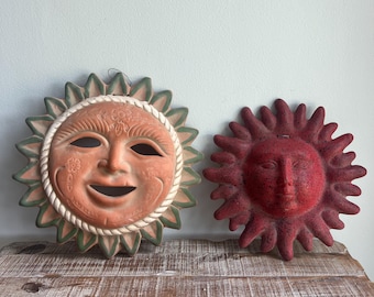 May include: Two sun-shaped wall plaques. The larger plaque is terracotta-colored with green-tipped rays, a rope-like border, and floral carvings. The smaller plaque is a solid, textured deep red with rounded rays and a serene facial expression.