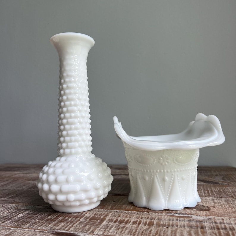 May include: A white milk glass vase with a hobnail texture and a narrow neck alongside a white milk glass hat-shaped bowl featuring a decorative embossed pattern with a scalloped rim. Both items are opaque and have a smooth, glossy finish.