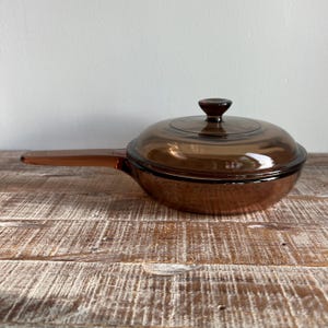 Op de afbeelding: Een doorschijnende, amberkleurige glazen koekenpan met bijpassend deksel. Het kookgerei is voorzien van een lange, geïntegreerde handgreep aan de zijkant en een ronde knop in het midden van het deksel. Het glas heeft een gladde, gepolijste afwerking.