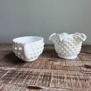 May include: Two pieces of white milk glass with a hobnail texture. The left piece is a small bowl with a single handle and a scalloped rim. The right piece is a small creamer or open sugar bowl featuring a ruffled, crimped edge and a hobnail pattern.