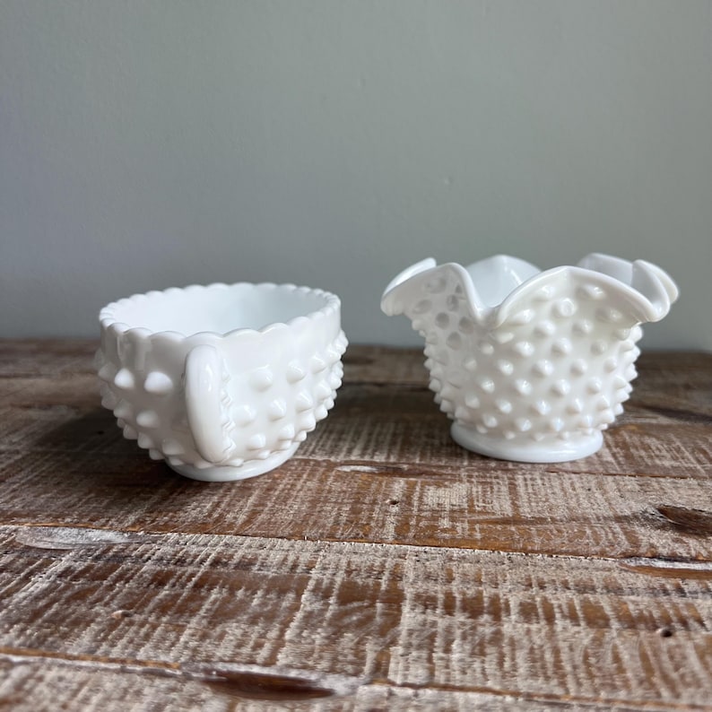May include: Two pieces of white milk glass with a hobnail texture. The left piece is a small bowl with a single handle and a scalloped rim. The right piece is a small creamer or open sugar bowl featuring a ruffled, crimped edge and a hobnail pattern.