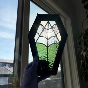 May include: A stained glass coffin-shaped suncatcher with a spiderweb design. The suncatcher features a black frame, clear and green textured glass, and is held up to a window. The spiderweb is made of clear glass.