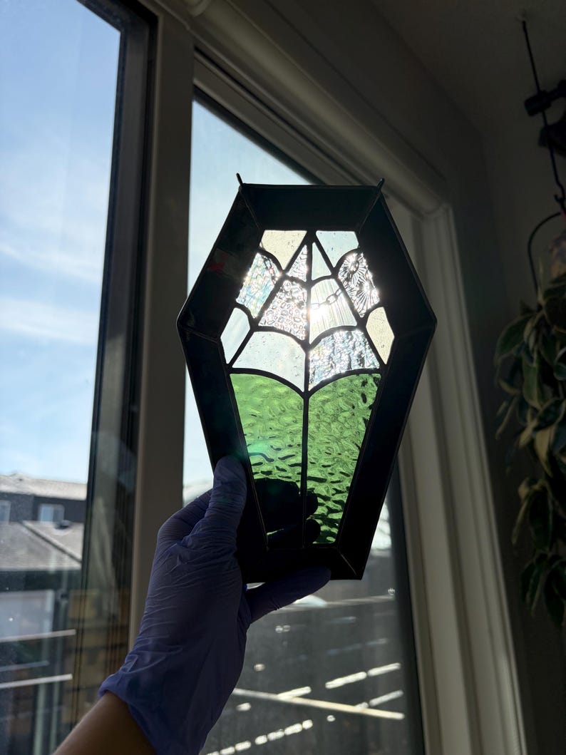 May include: A stained glass coffin-shaped suncatcher with a spiderweb design. The suncatcher features a black frame, clear and green textured glass, and is held up to a window. The spiderweb is made of clear glass.