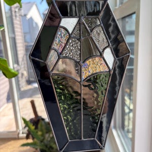 May include: A stained glass coffin-shaped suncatcher with a silver chain for hanging. The design features black and iridescent glass panels, creating a unique and decorative piece. The suncatcher is displayed near a window, catching the light.