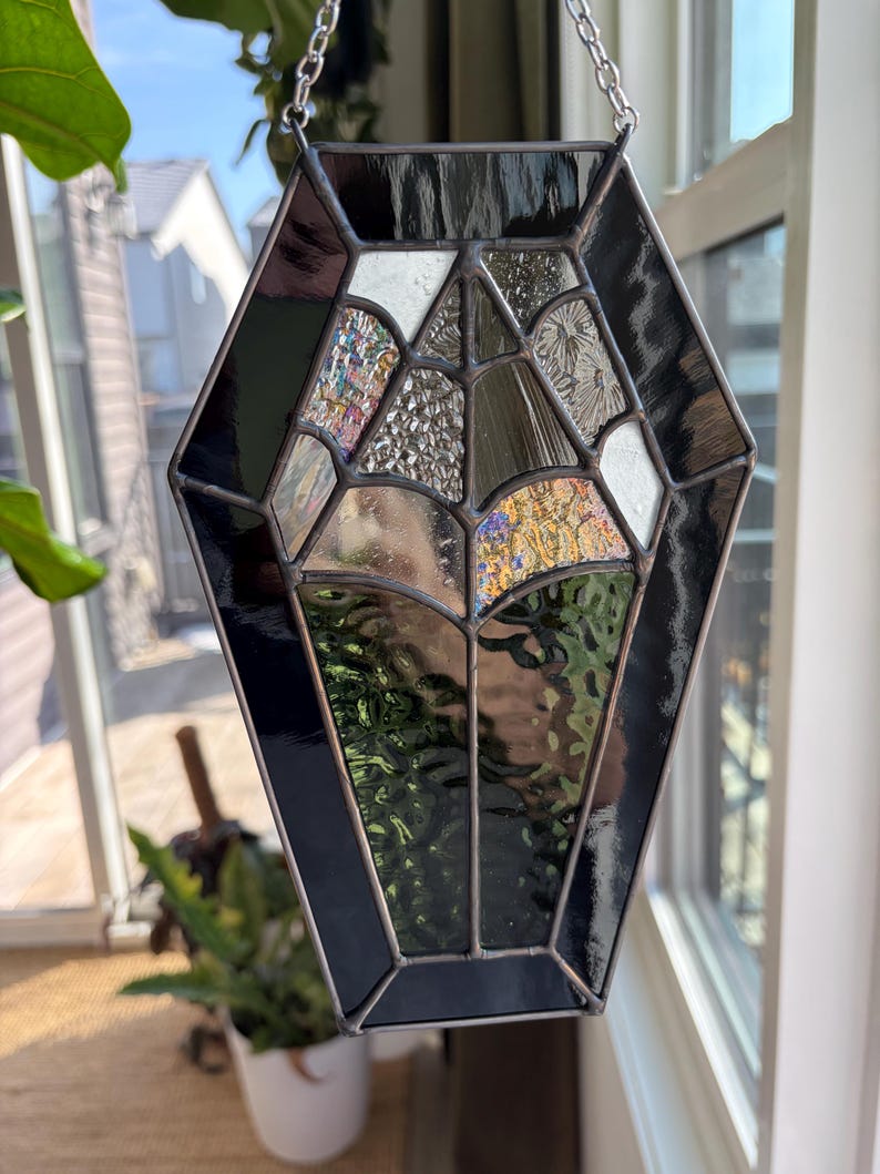 May include: A stained glass coffin-shaped suncatcher with a silver chain for hanging. The design features black and iridescent glass panels, creating a unique and decorative piece. The suncatcher is displayed near a window, catching the light.