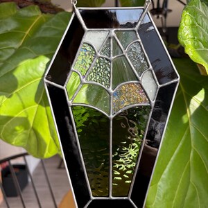 May include: A stained glass suncatcher in a coffin shape, featuring a black border and a web-like design. The glass pieces are in shades of green, white, and gold, with a silver chain for hanging. The suncatcher is displayed against a backdrop of green leaves.