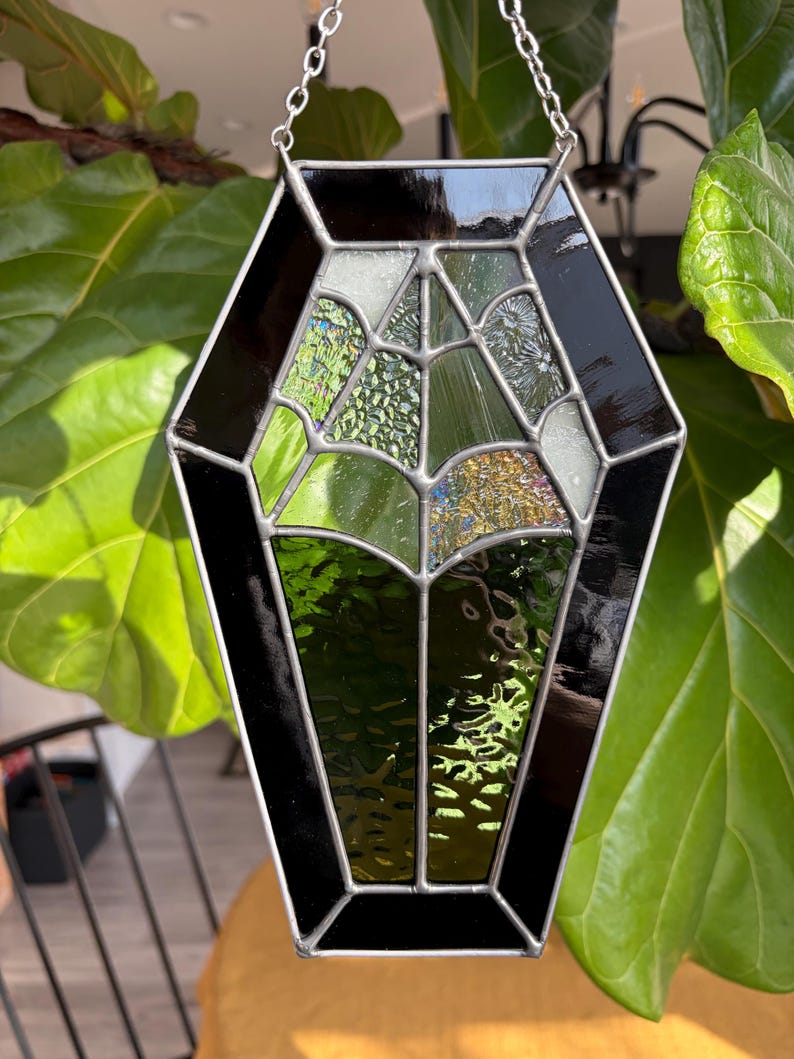 May include: A stained glass suncatcher in a coffin shape, featuring a black border and a web-like design. The glass pieces are in shades of green, white, and gold, with a silver chain for hanging. The suncatcher is displayed against a backdrop of green leaves.