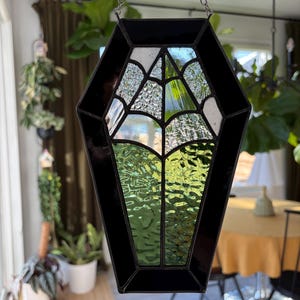 May include: A black coffin-shaped stained glass suncatcher with a spiderweb design. The glass is a mix of clear, white, and green, with a black border. It hangs from a silver chain, perfect for Halloween or gothic decor.