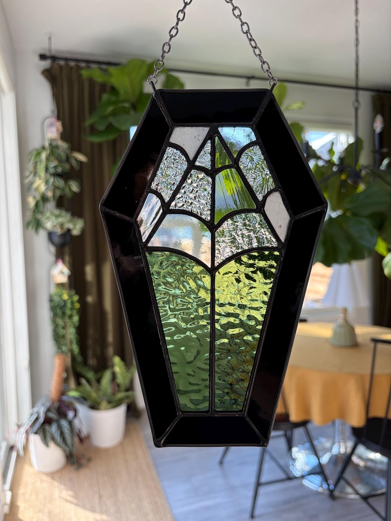 May include: A black coffin-shaped stained glass suncatcher with a spiderweb design. The glass is a mix of clear, white, and green, with a black border. It hangs from a silver chain, perfect for Halloween or gothic decor.