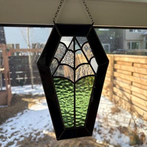 May include: A stained glass coffin-shaped suncatcher with a black border and a chain for hanging. The glass features a spiderweb design, clear textured glass, and green textured glass. The suncatcher is displayed in front of a window.