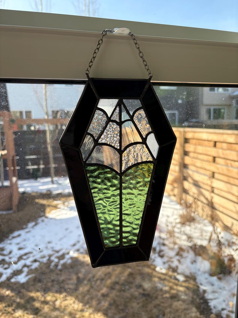 May include: A stained glass coffin-shaped suncatcher with a black border and a chain for hanging. The glass features a spiderweb design, clear textured glass, and green textured glass. The suncatcher is displayed in front of a window.