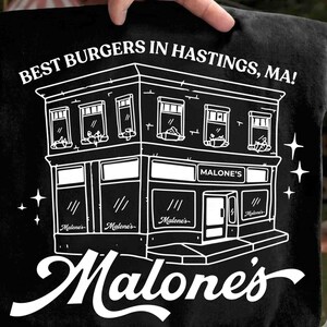 May include: Black t-shirt featuring a white line drawing of a two-story building with the text "BEST BURGERS IN HASTINGS, MA!" and "Malone's". The building has windows and doors, with the name "Malone's" repeated.