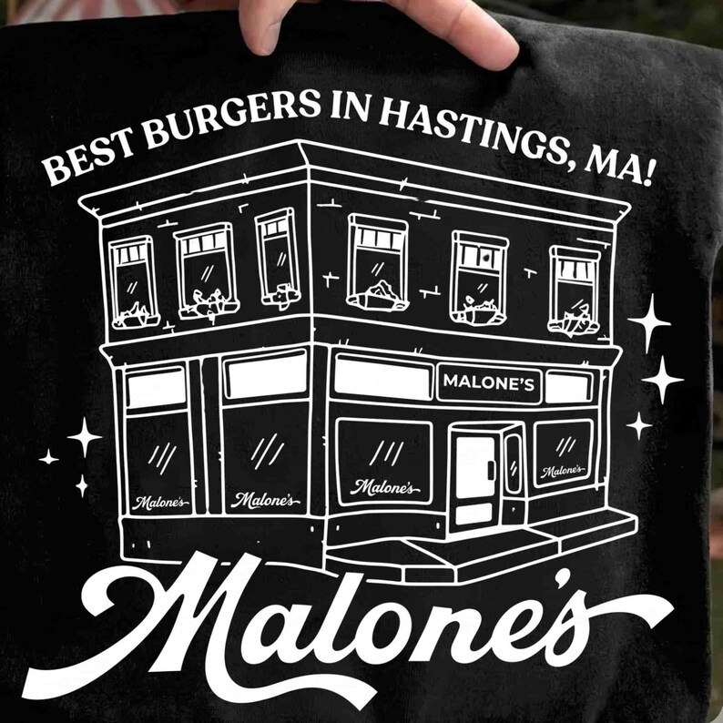 May include: Black t-shirt featuring a white line drawing of a two-story building with the text "BEST BURGERS IN HASTINGS, MA!" and "Malone's". The building has windows and doors, with the name "Malone's" repeated.